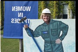 Open House 1992; woman posing in face in hole board in front of Women in Trades Banner