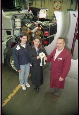 Open House 1998; group of three holding white and green ribbon in front of semi truck with engine...