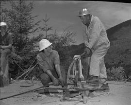 Logging, 1967; a man holdings a large metal cable and another man using logging equipment