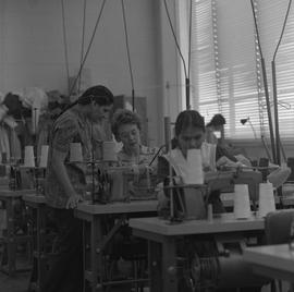 Sewing, 1971; woman sitting at a sewing machine talking to another woman ; people working in back...