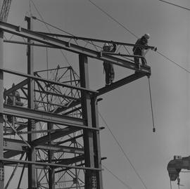 Structural steel, 1971; two workers standing on a side beam guiding a large piece of steel