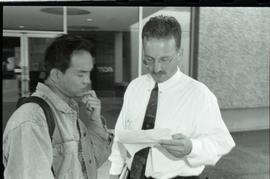 Communications Relations Opening Day 1991; man in tie holding paper beside student in jean jacket...
