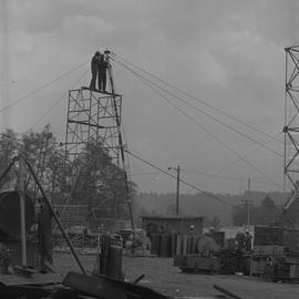 Structural steel, 1971; two men standing at the top of scaffolding holding onto a tall pole; peop...